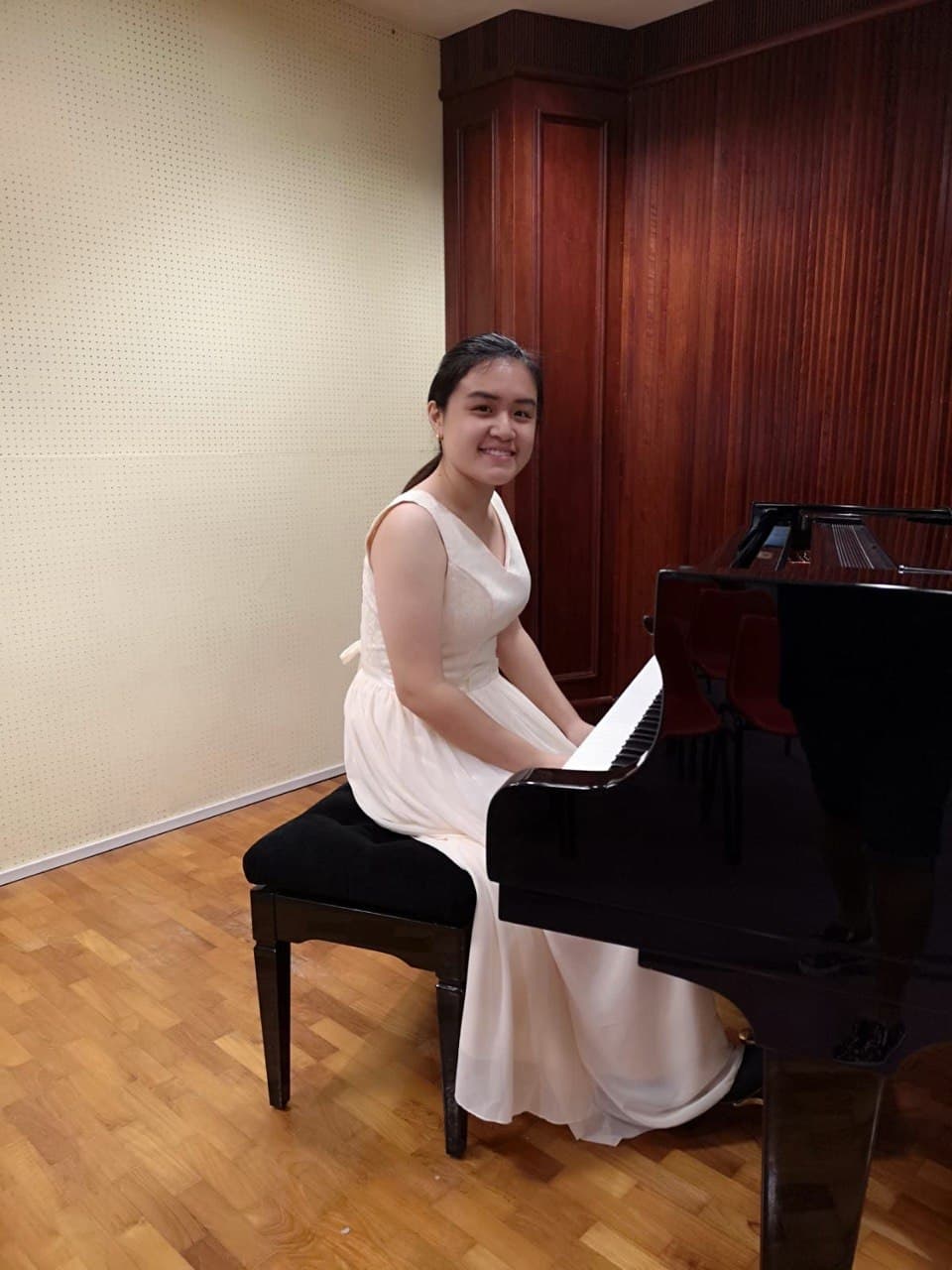 Smiling young woman in a white sleeveless dress sits at a black grand piano.