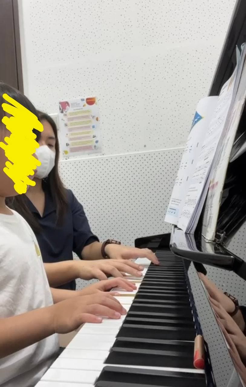 A masked teacher and young student play the piano together during a music lesson.