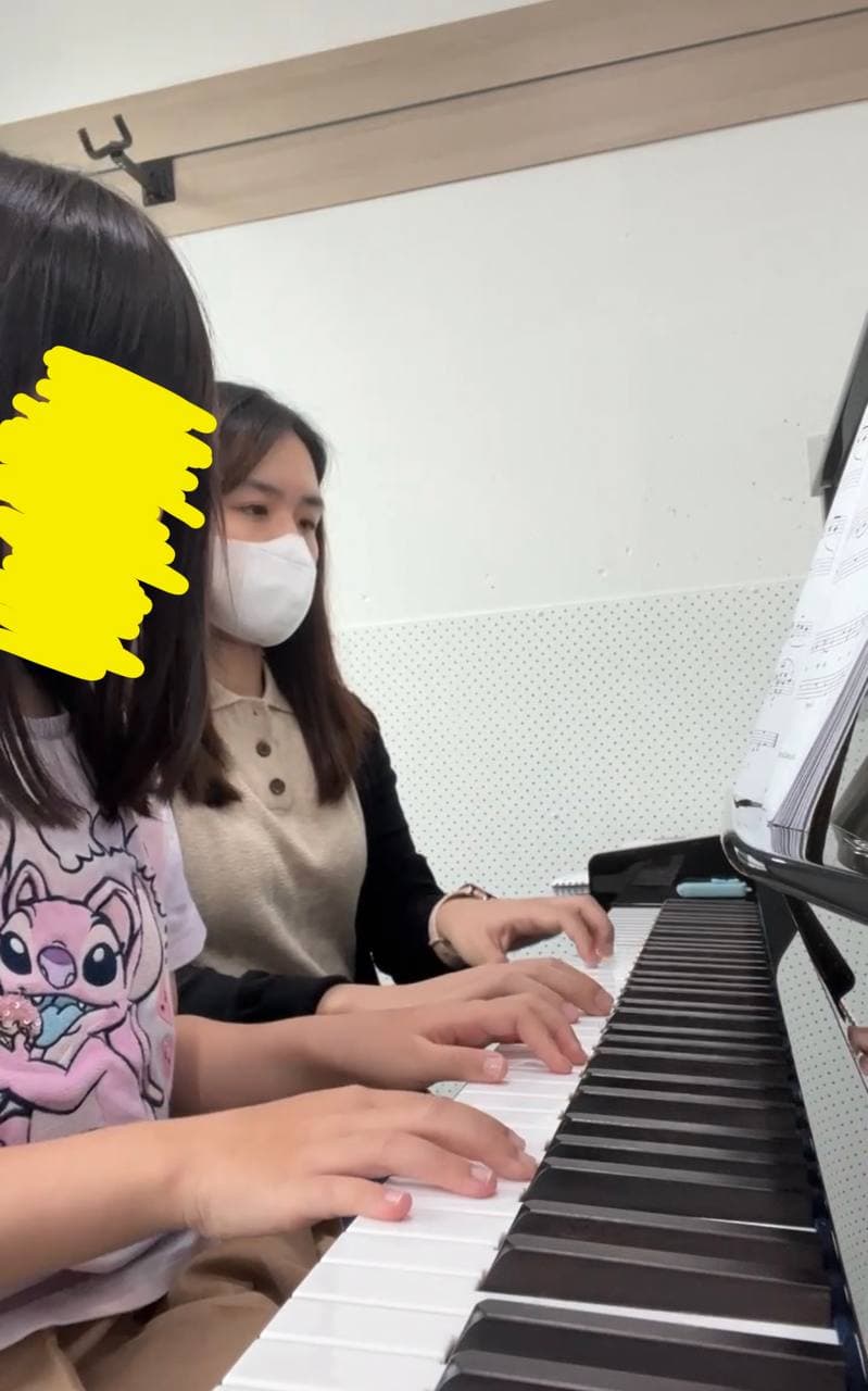 A masked teacher and a young student play the piano together during a music lesson.